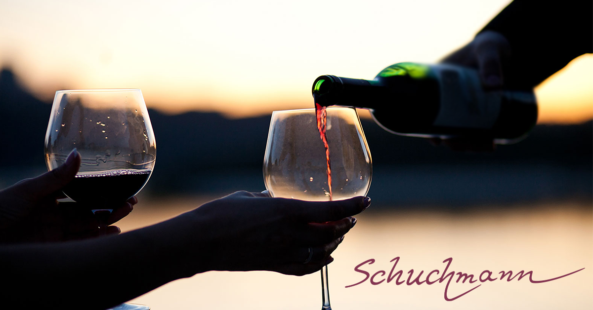 Zwei Hände halten Weingläser bei Sonnenuntergang, die mit Rotwein gefüllt werden, mit dem Schriftzug 'Schuchmann' in eleganter Schrift