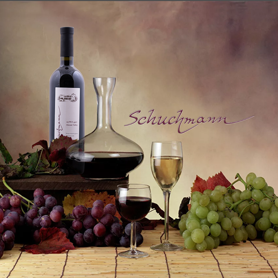 Schuchmann-Weinflasche mit Dekanter, Rot- und Weißweinglas, umgeben von bunten Trauben."